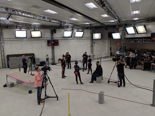 three people with motion capture suits on and a ot of other people standing with cameras surrounding them, the studio is also full of other props and lights